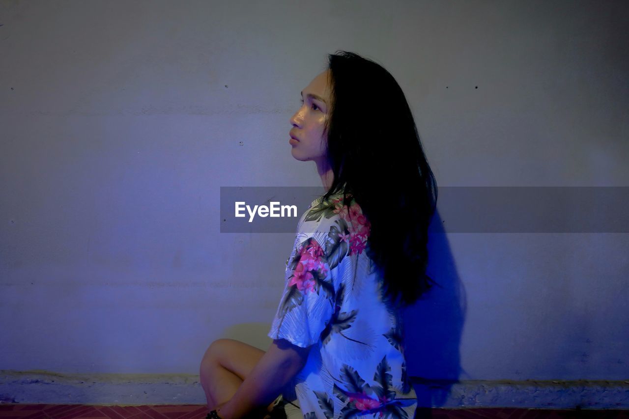 Side view of young woman looking away while sitting against wall at home