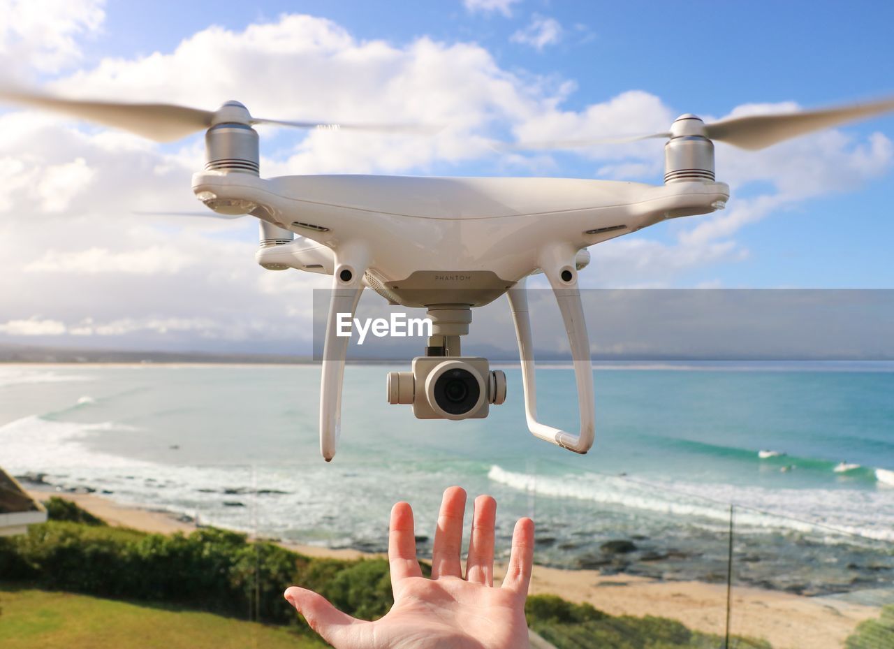 Cropped hand gesturing towards drone flying at beach against sky