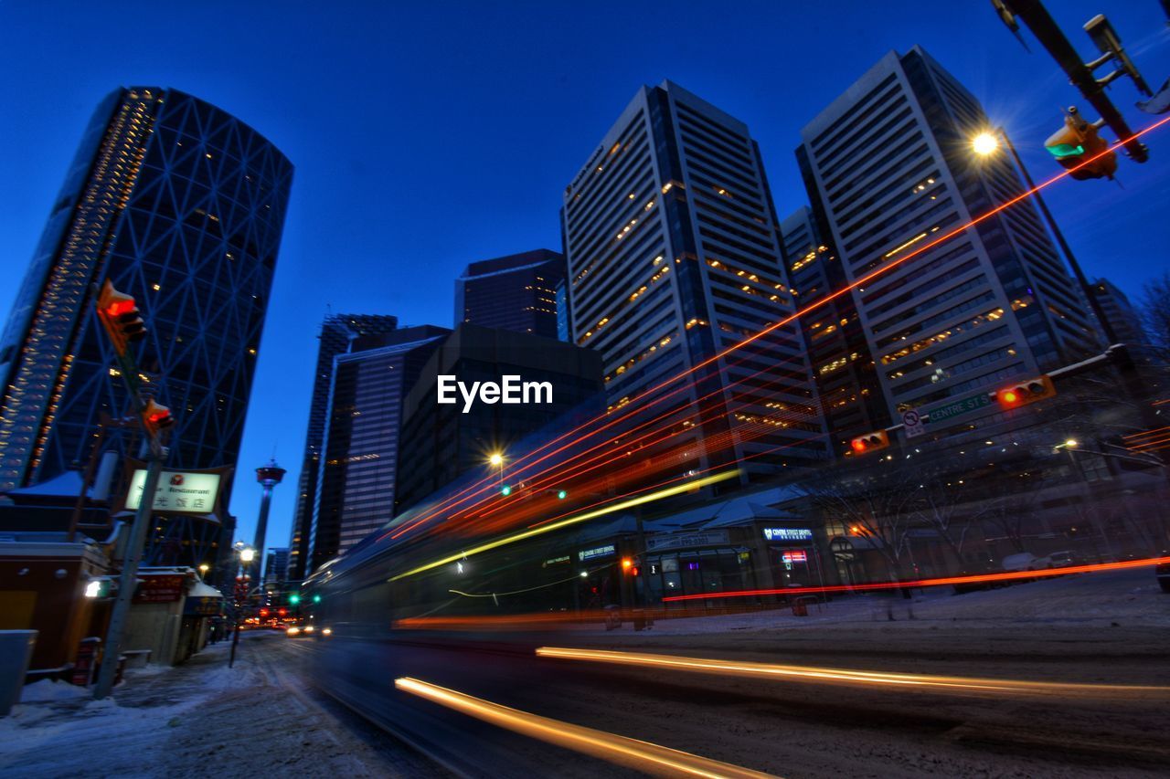 LIGHT TRAILS ON ROAD BY BUILDINGS AGAINST SKY AT NIGHT