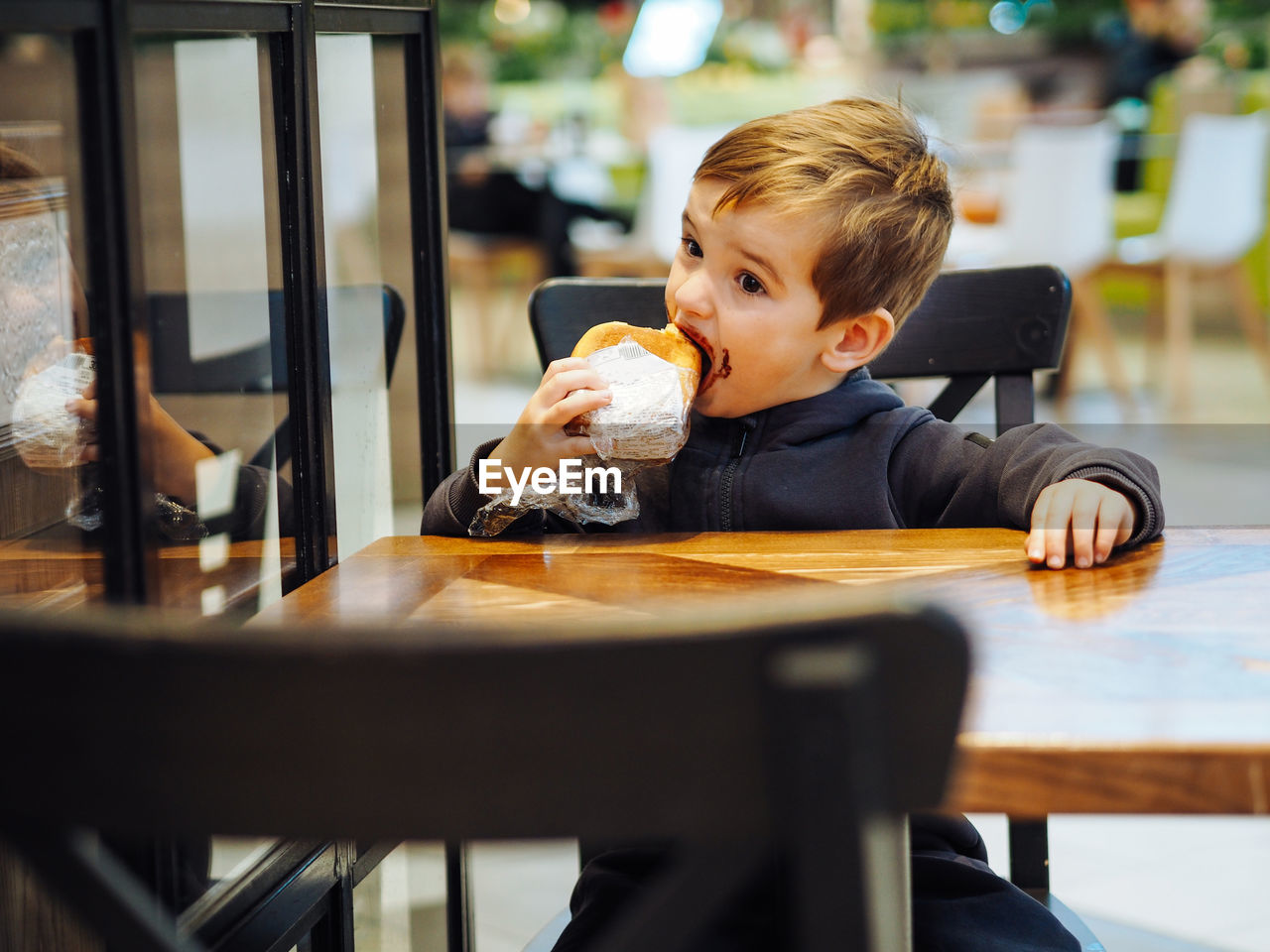 Toddler boy in casual clothes eating a chocolate bun in a food court there is chocolate on his face