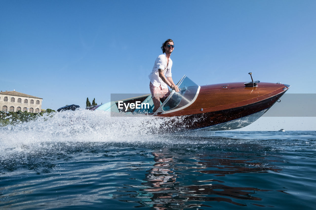 Low angle view of young man riding speedboat on sea against clear blue sky