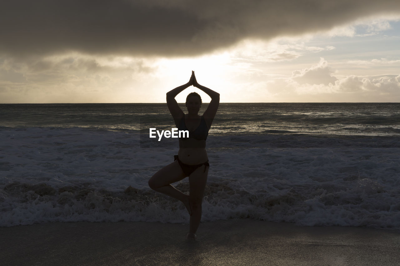 sea, sky, water, beach, one person, exercising, yoga, land, horizon over water, adult, spirituality, horizon, lifestyles, nature, sports, wellbeing, meditating, religion, women, beauty in nature, zen-like, arm, stretching, leisure activity, sunset, physical fitness, full length, flexibility, limb, tranquility, relaxation, holiday, vitality, balance, arms raised, young adult, cloud, vacation, trip, tranquil scene, female, motion, standing, human limb, idyllic, scenics - nature, sand, practicing, silhouette, environment, rear view, harmony, ocean, clothing, sunlight, outdoors, solitude, wave, posture, strength, dusk, martial arts