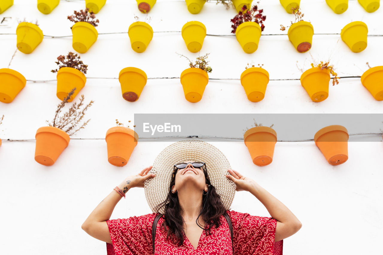 Cheerful female tourist in straw hat standing near wall with assorted plants in pots in rhodes on white background