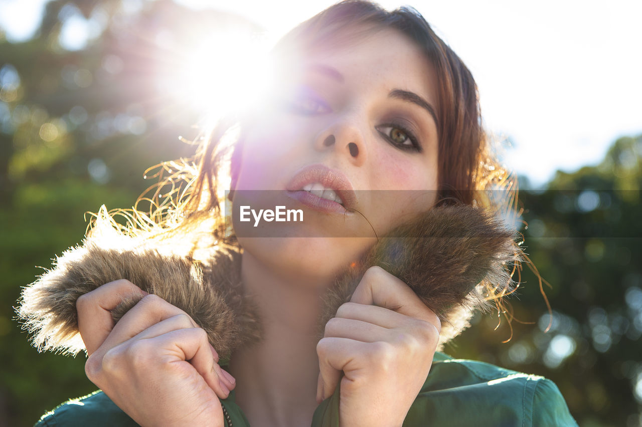 Close-up portrait of young woman in warm clothing against sky at park during sunny day