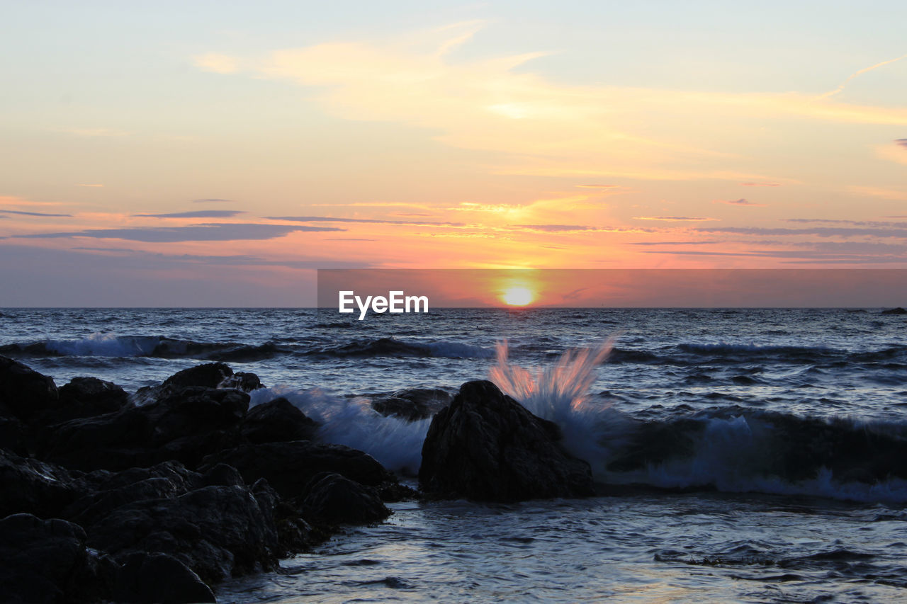 View of sea waves splashing on rocks against sky during sunset