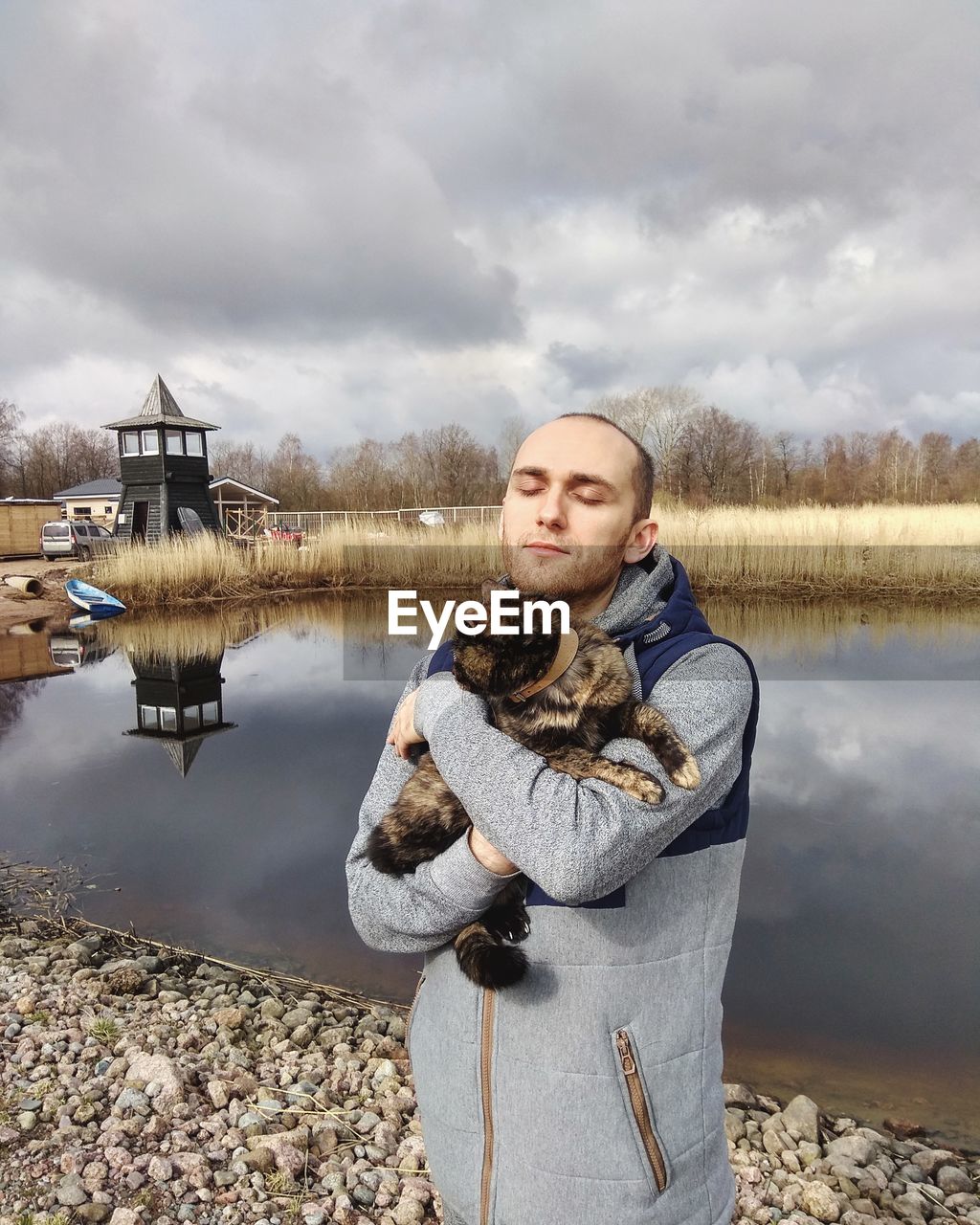 Man with cat standing by lake against cloudy sky