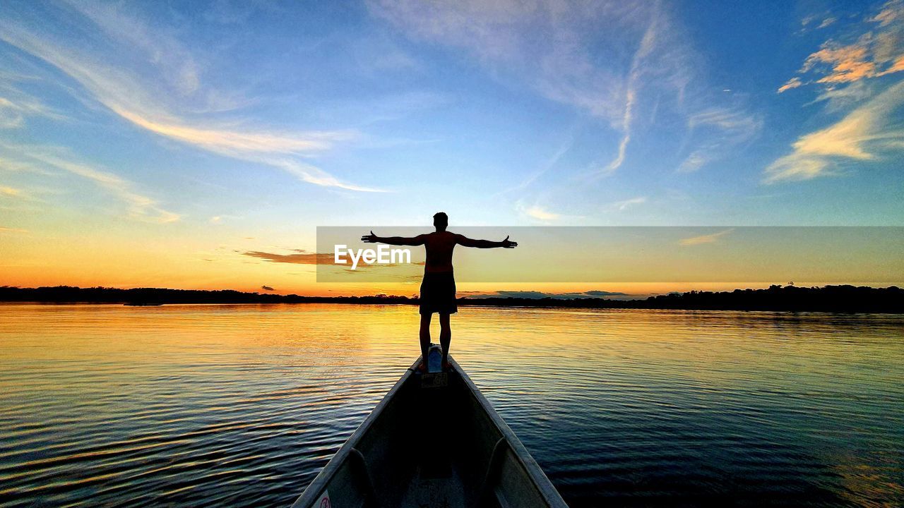 Silhouette man standing in boat by lake against sky during sunset
