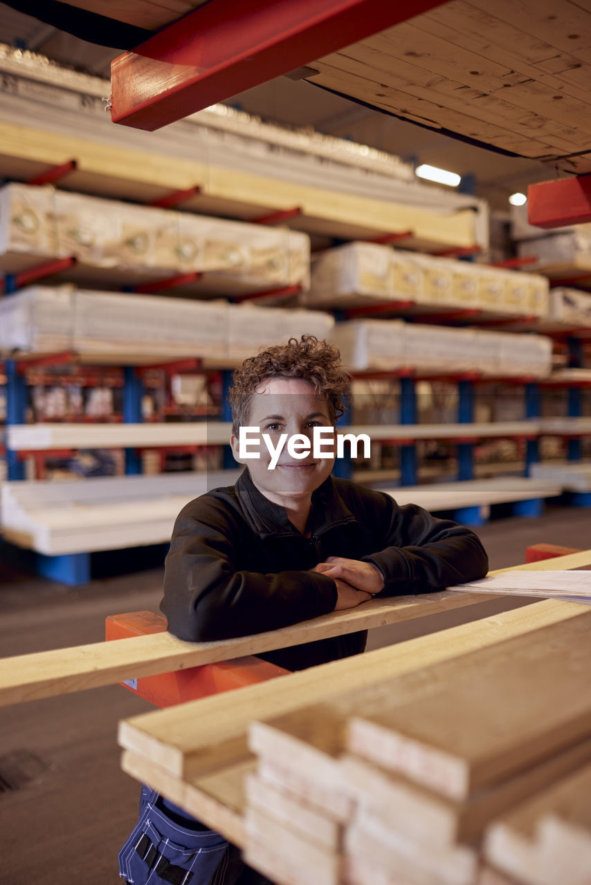 Portrait of smiling mature female worker leaning on plank at lumber industry