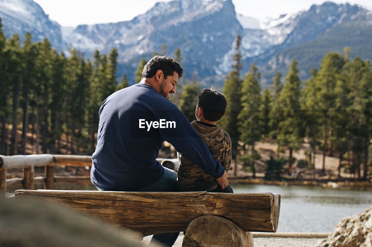 Father and son sitting by lake against mountain