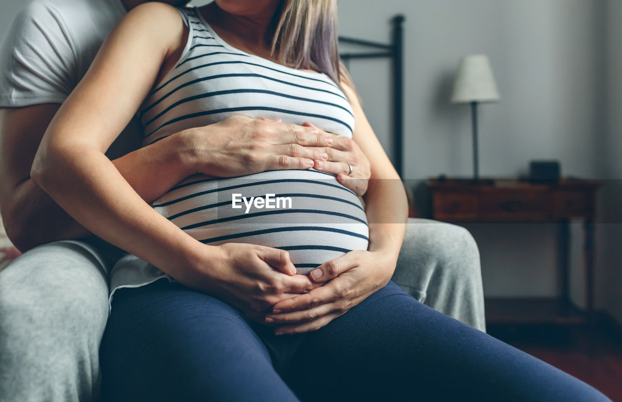 Midsection of pregnant woman standing with man sitting at home