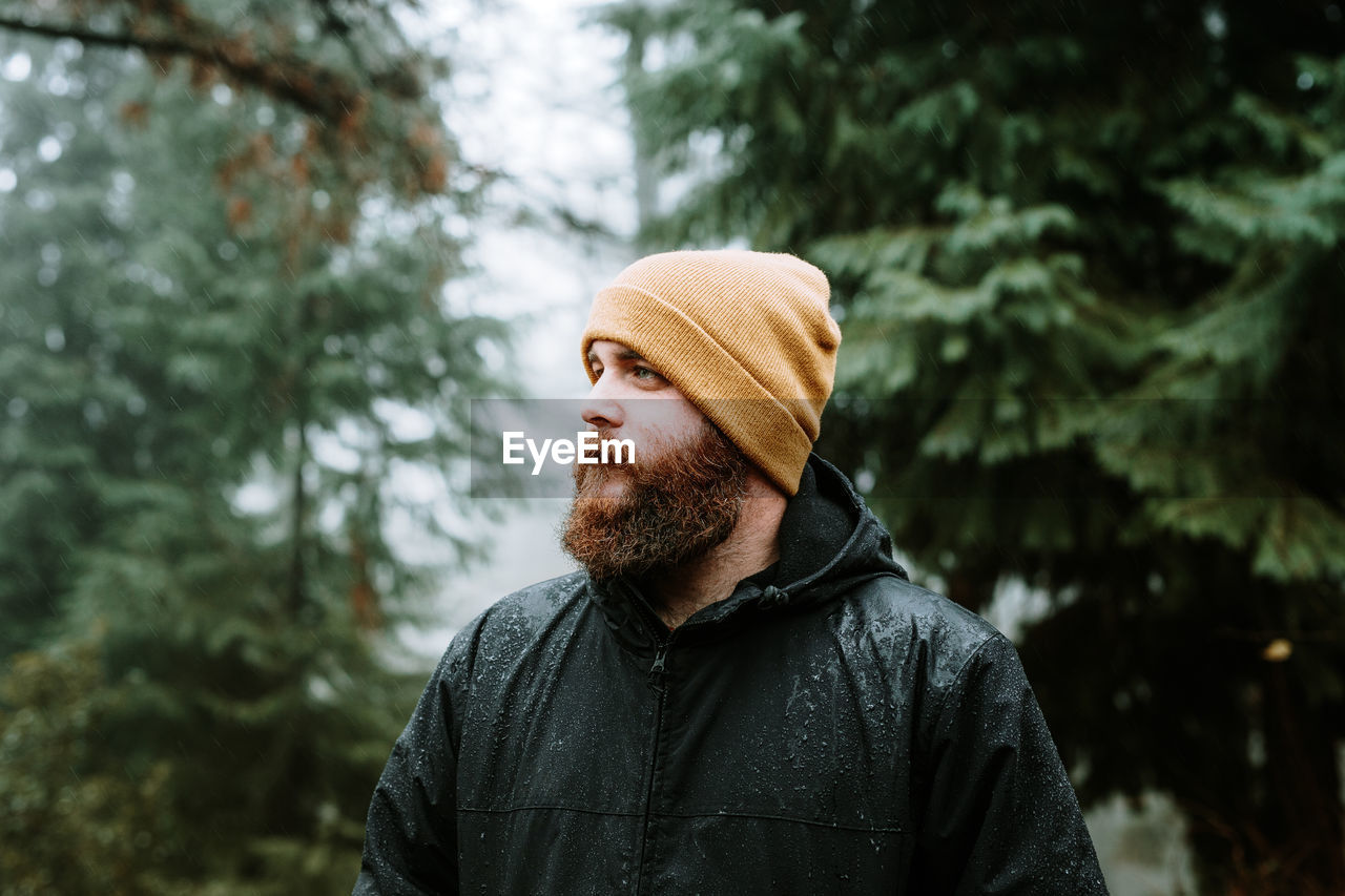 Bearded guy in coat and hat near fir trees