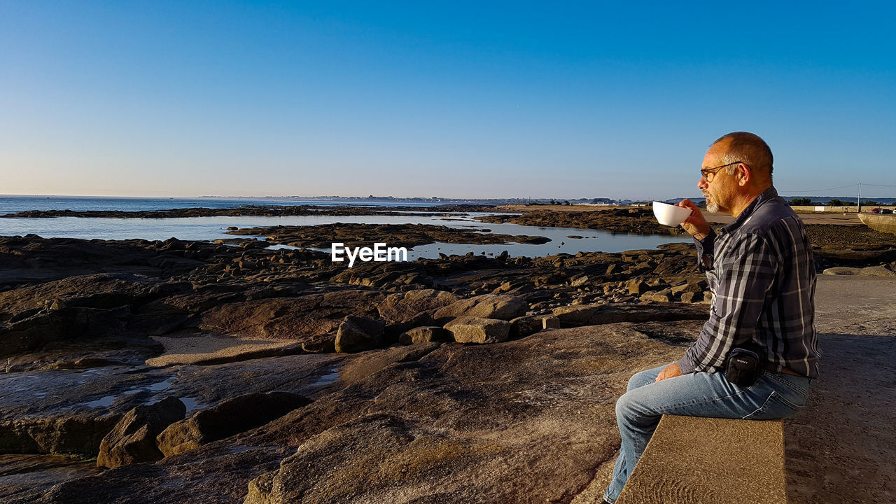 Man drinking coffee while sitting on rock at beach against sky