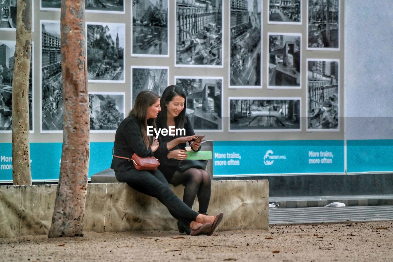 MEN SITTING ON MAN USING MOBILE PHONE WHILE LOOKING AT CAMERA