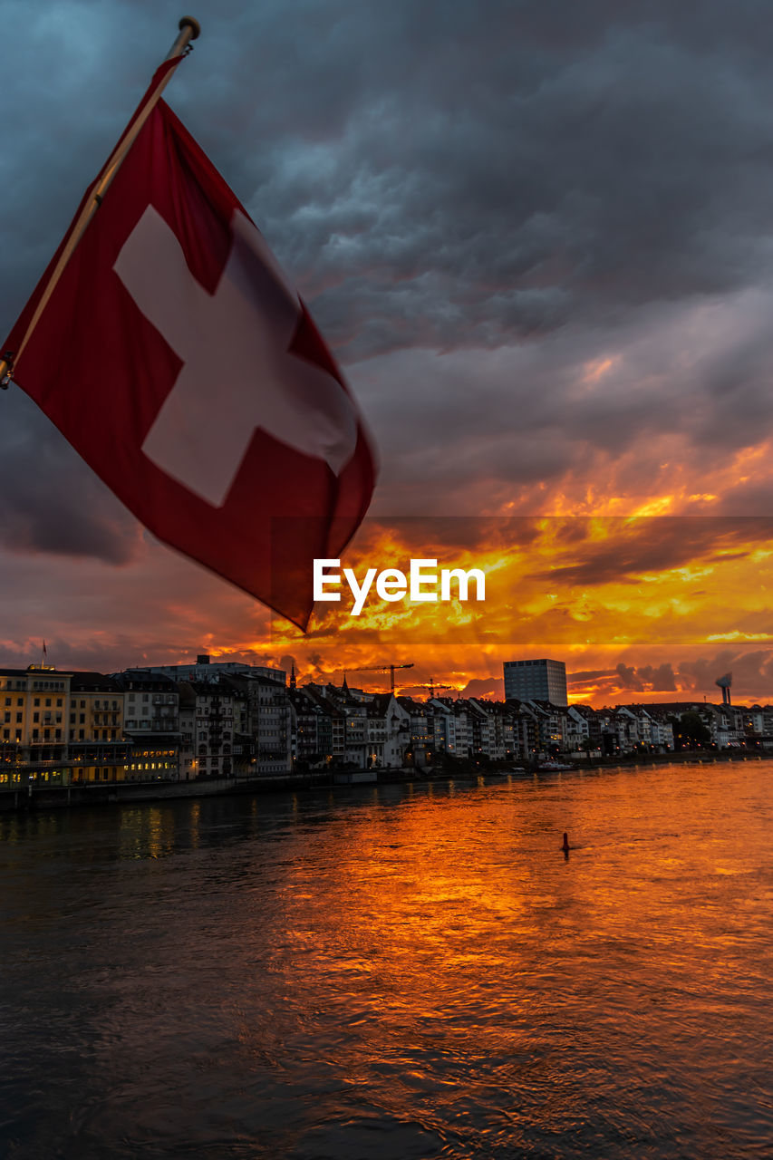 FLAG BY RIVER AND BUILDINGS AGAINST ORANGE SKY