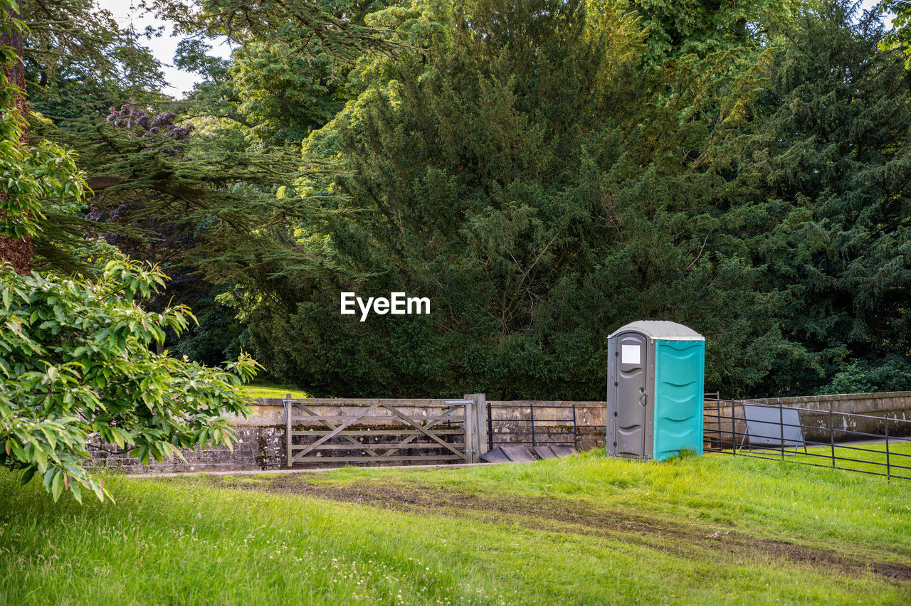 A plastic portable toilet next to muddy track in field at outdoor event and surrounded by tall trees