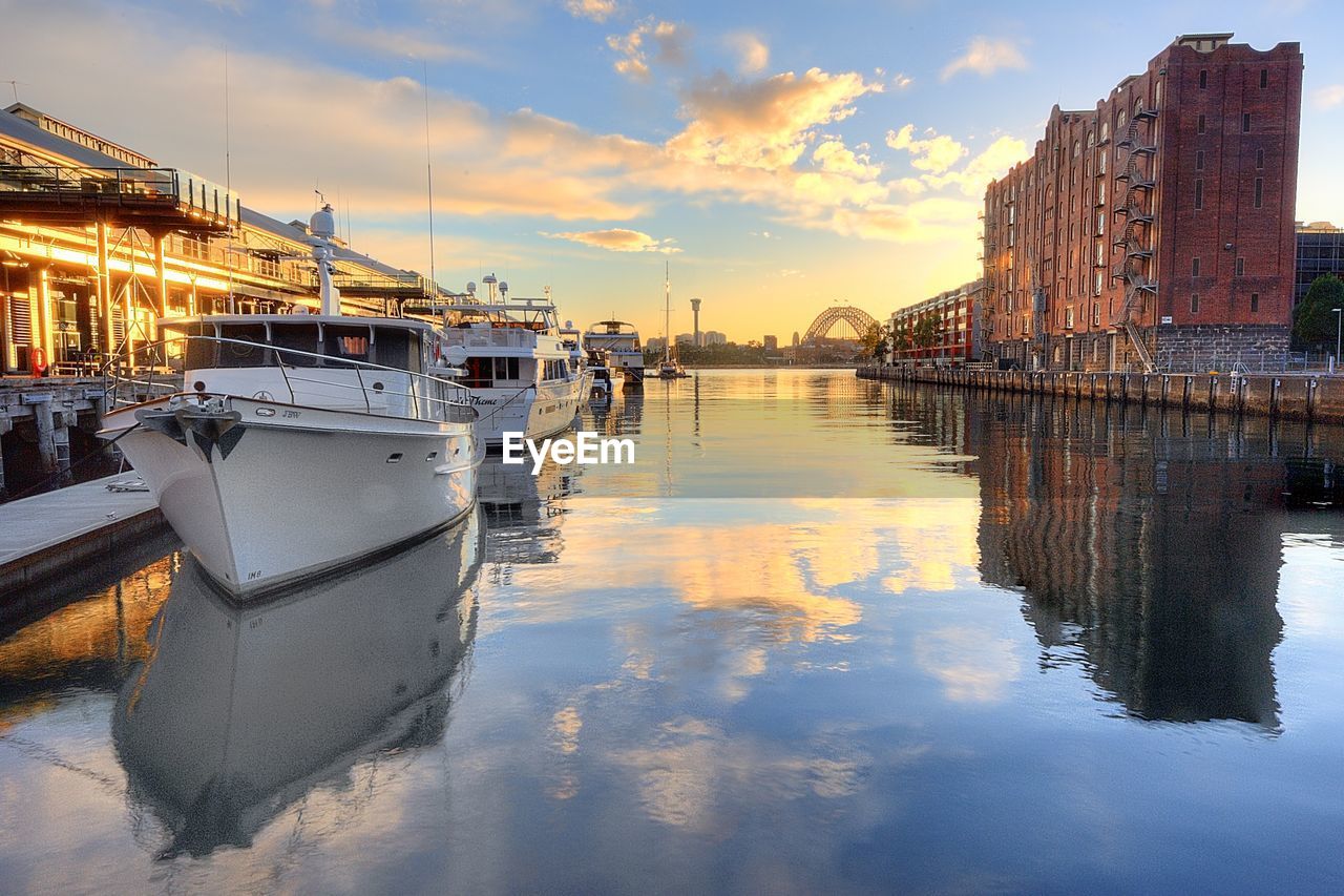 Boats moored at harbor by building against sky during sunset