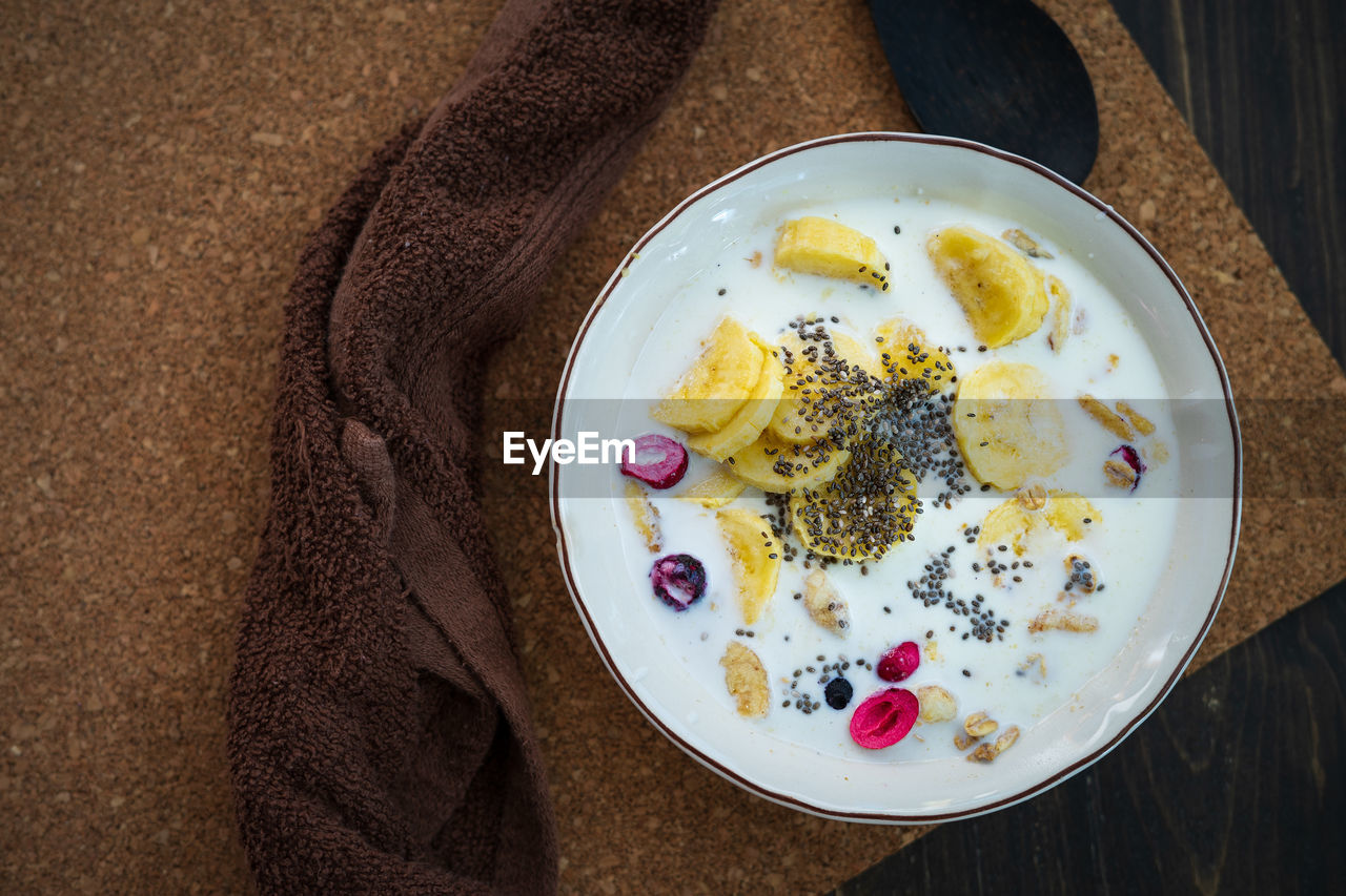 Fresh granola, banana with milk in a white bowl on brown background. sprinkled with chia seeds.