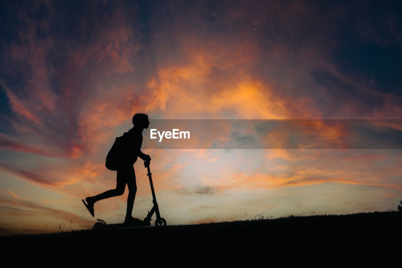 Silhouette of a child riding a scooter against dramatic sunset sky