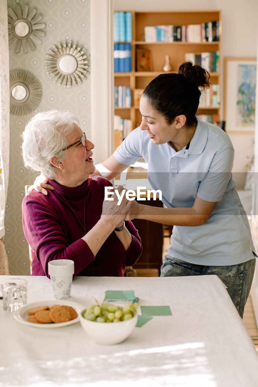 Smiling retired senior woman holding hands of young female volunteer while talking in nursing home