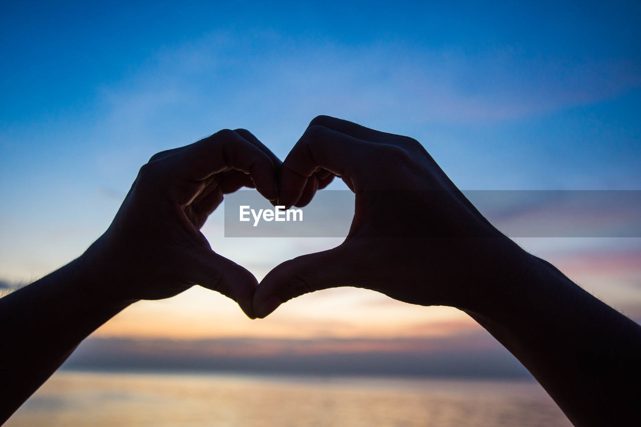 Cropped hands forming heart shape against sea during sunset