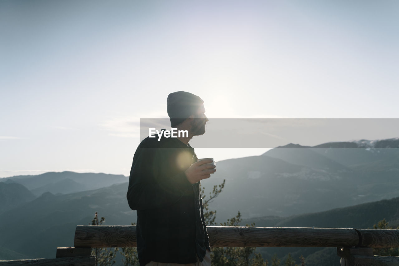 Side view of man looking at mountains against sky having a cup of coffee