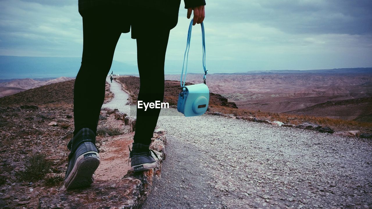 Low section of woman with purse walking on footpath against cloudy sky