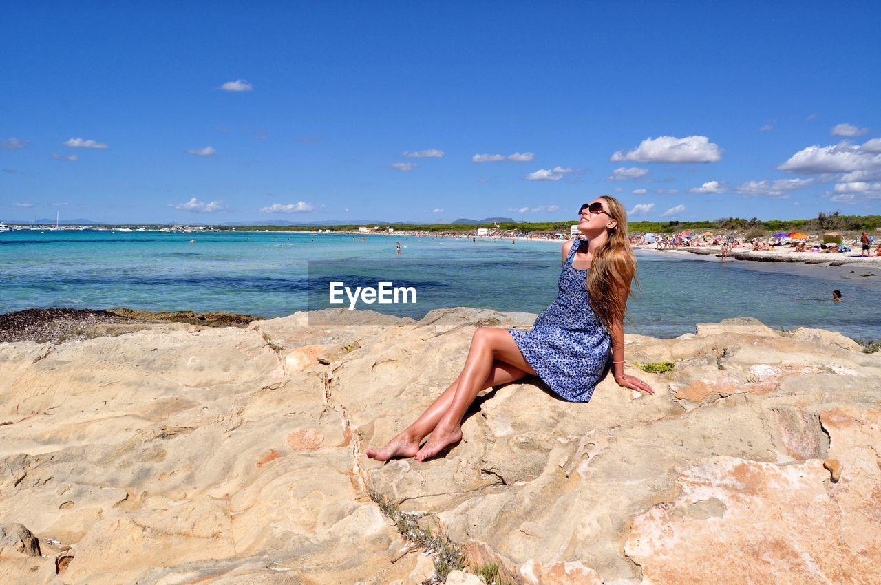 Woman sitting on rock at beach against blue sky