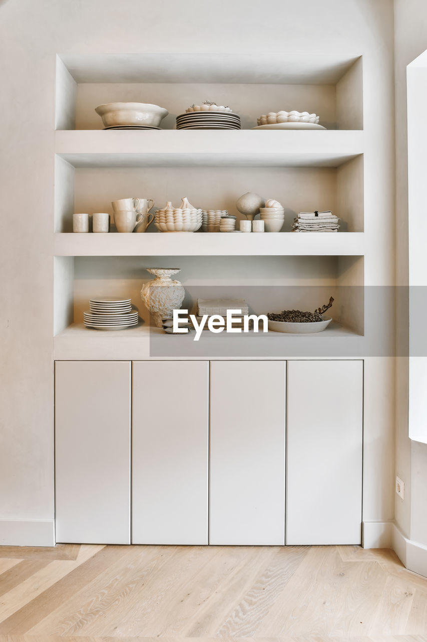 Elegant ceramic plates cups and vases arranged on built in shelves in minimalist style apartment with wooden parquet floor in daylight