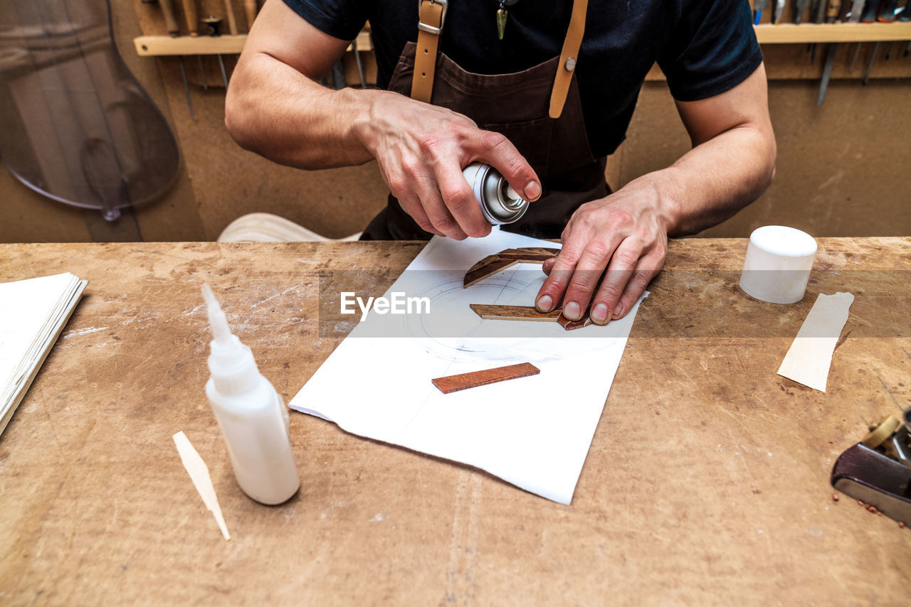 From above of crop anonymous male artisan applying adhesive spray on wooden piece on paper sheet in workroom