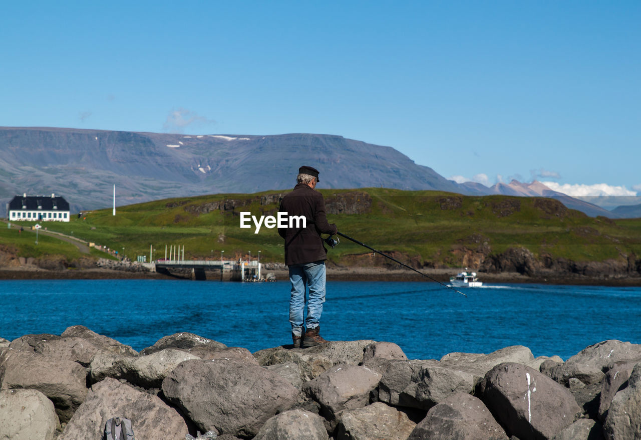 Full length rear view of man fishing while standing on rocks at lakeshore