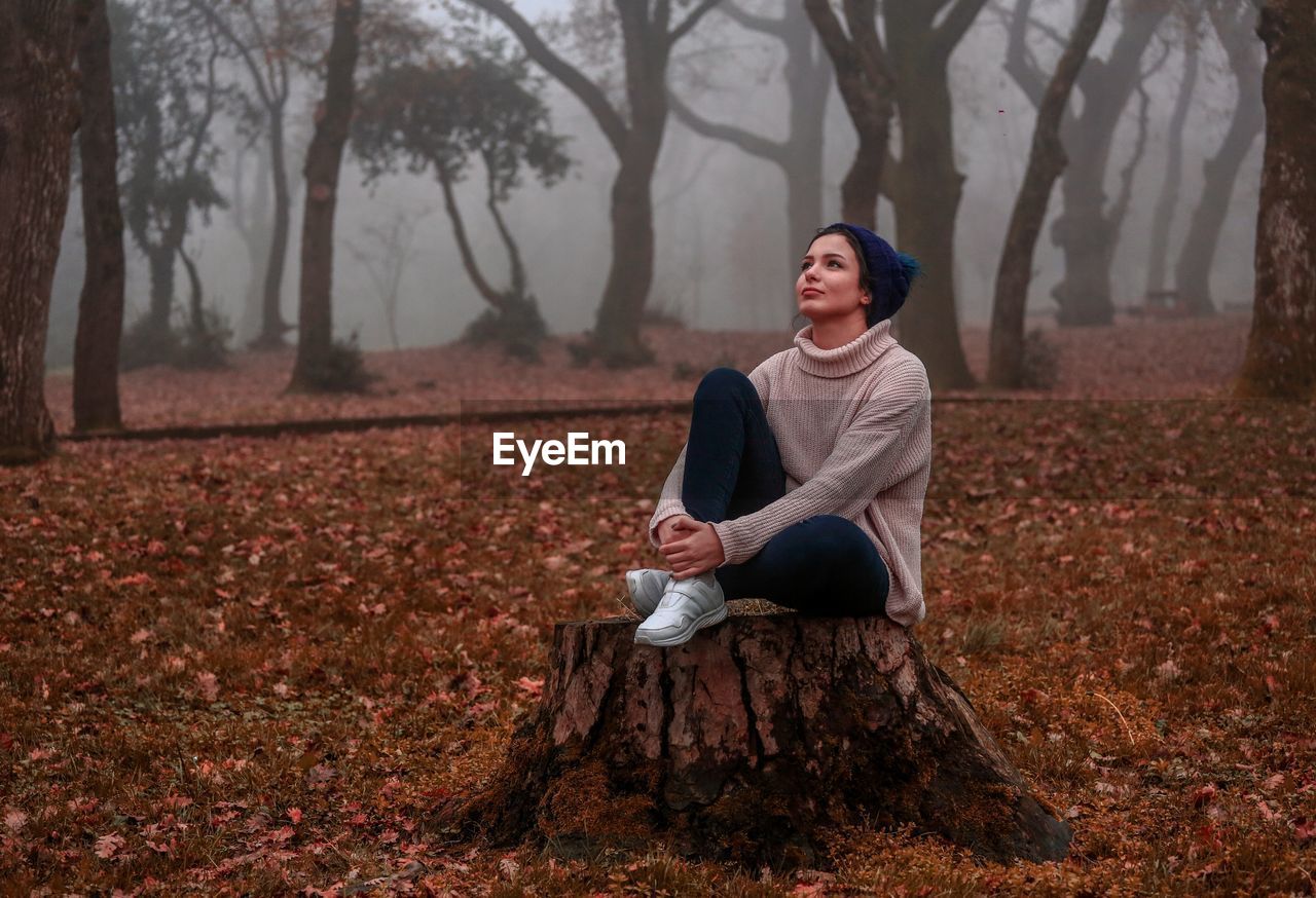 Full length of thoughtful young woman sitting on tree stump in forest during autumn