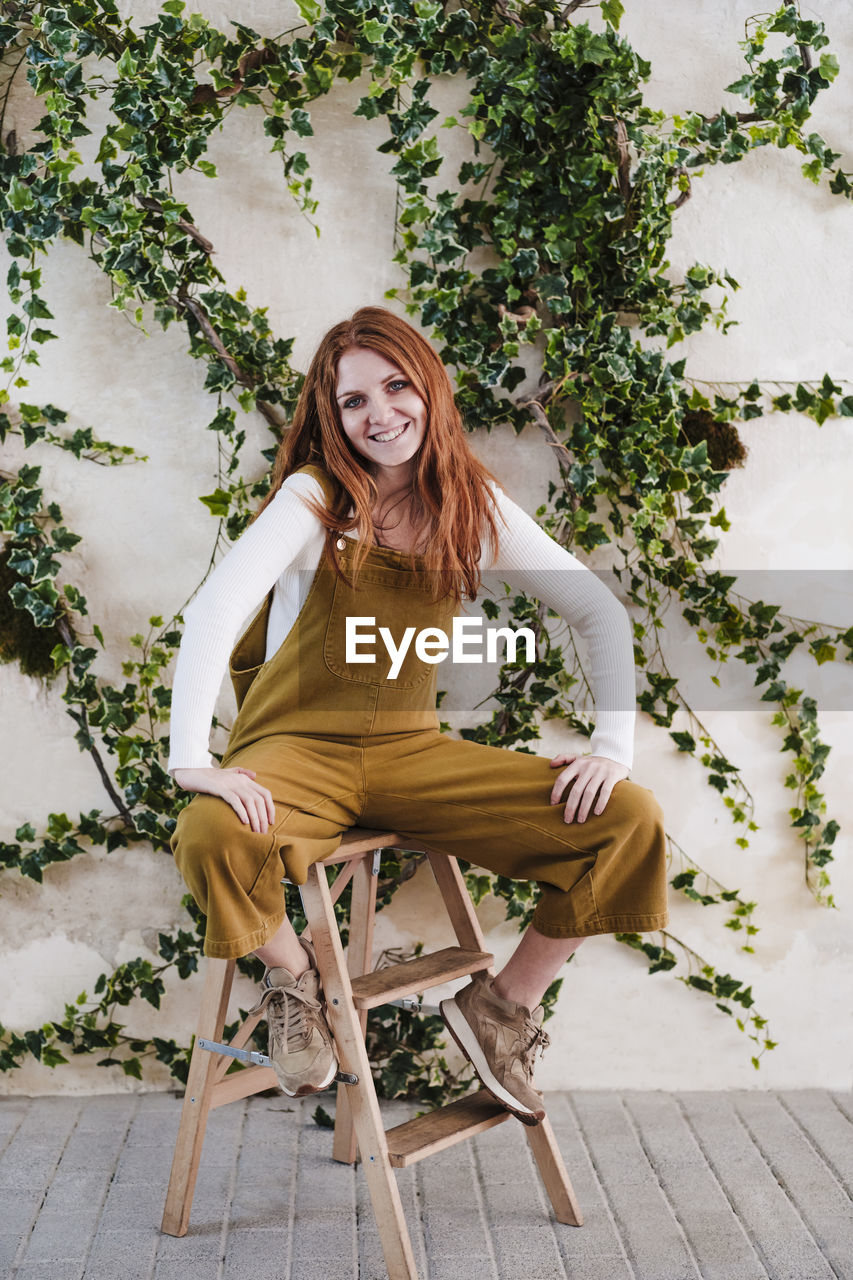 Smiling woman sitting on step ladder in front of vines