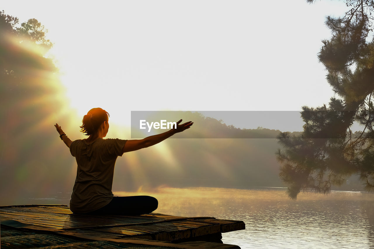 rear view of woman with arms raised standing by lake against sky during sunset