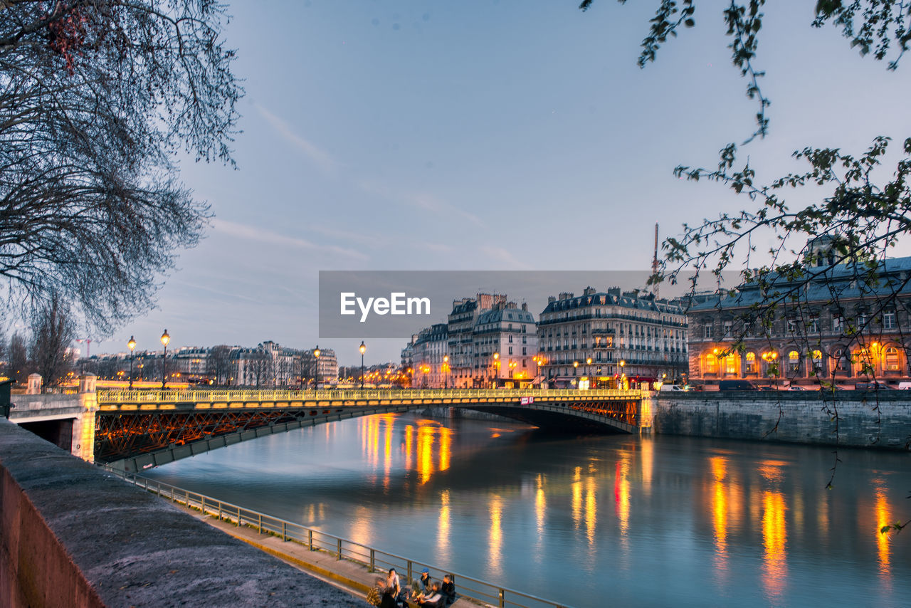 Bridge over river by illuminated city against sky