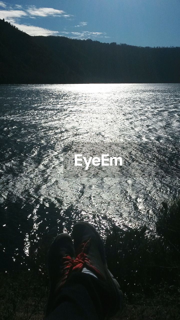 LOW SECTION OF MAN STANDING IN SEA AGAINST MOUNTAINS