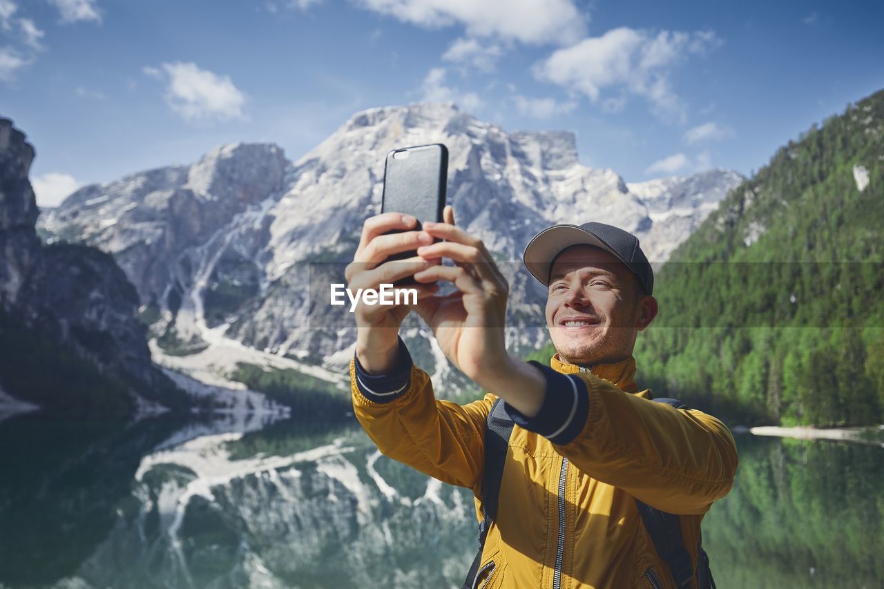 Smiling hiker taking selfie through mobile phone against lake and mountains