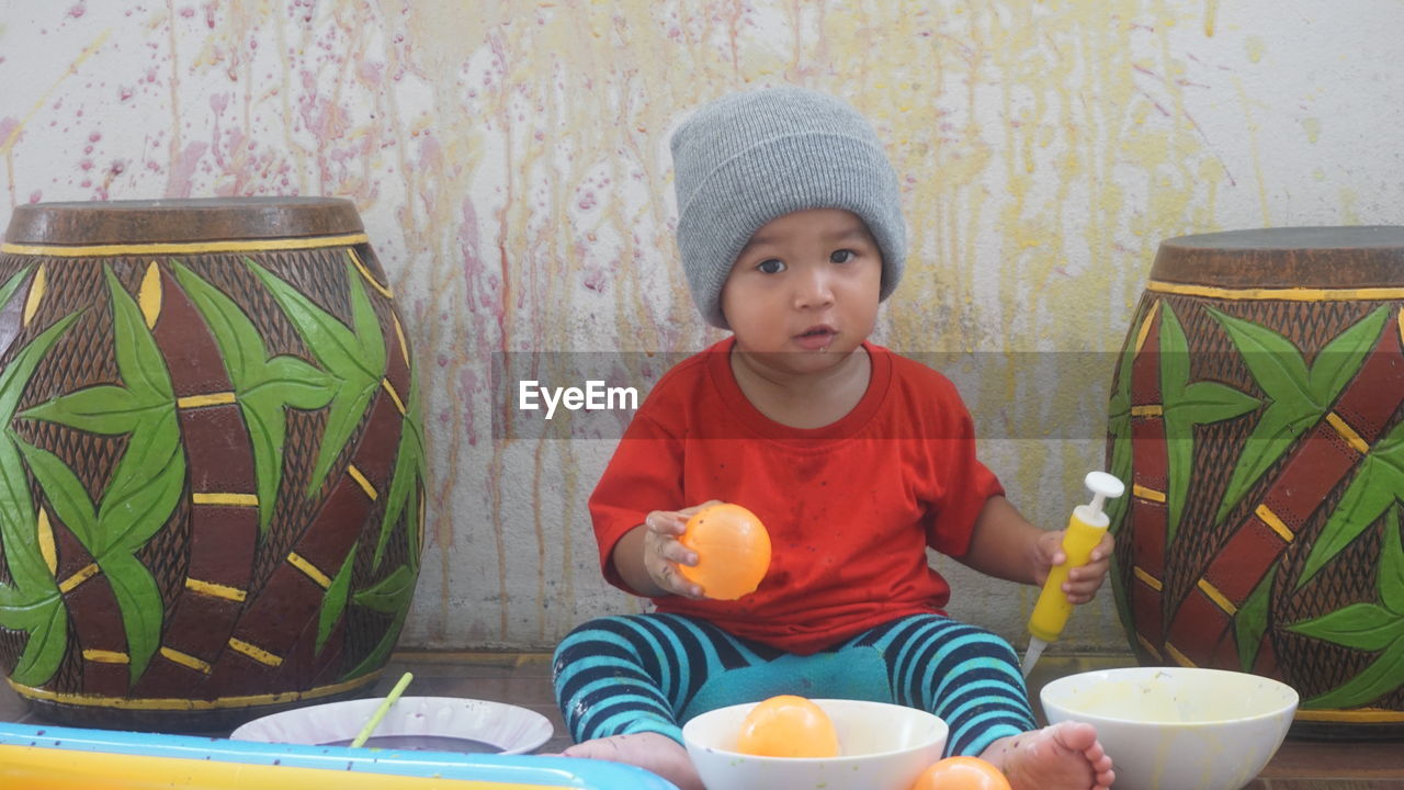 Portrait of cute baby boy holding water bombs while sitting against wall