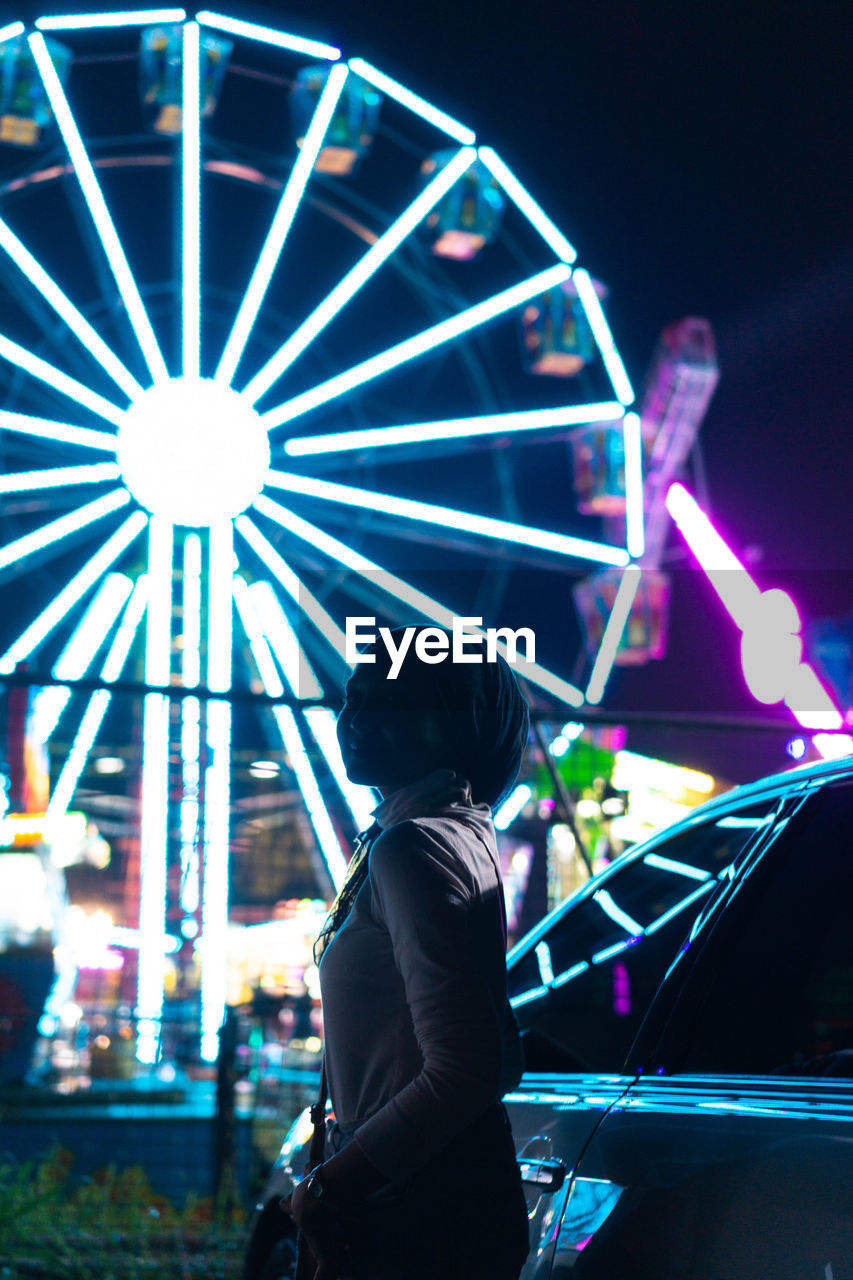 Side view of woman standing at amusement park ride at night