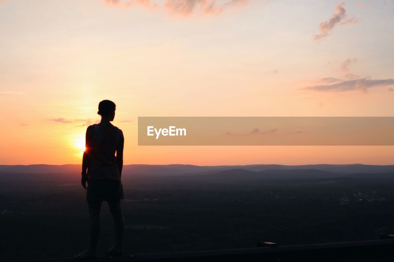 Rear view of silhouette boy standing by landscape against sky during sunset