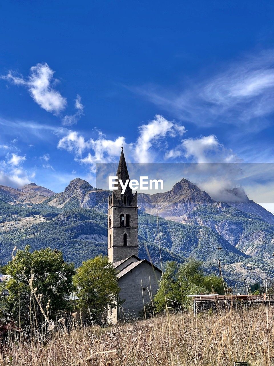 sky, architecture, mountain, place of worship, religion, built structure, mountain range, landscape, building, building exterior, belief, cloud, nature, scenics - nature, spirituality, travel destinations, plant, environment, history, beauty in nature, blue, the past, land, travel, no people, catholicism, tower, rural area, tree, tourism, grass, rural scene, outdoors, spire, non-urban scene, tranquility, day, tranquil scene, steeple, field, idyllic, old, mountain peak