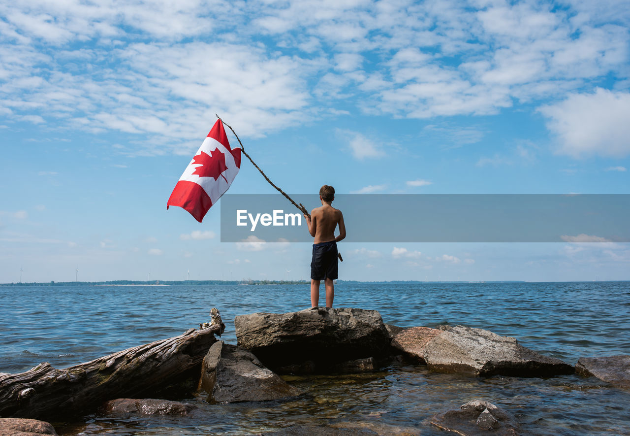 Boy holding canada flag in the air on shore of a lake on a summer day.