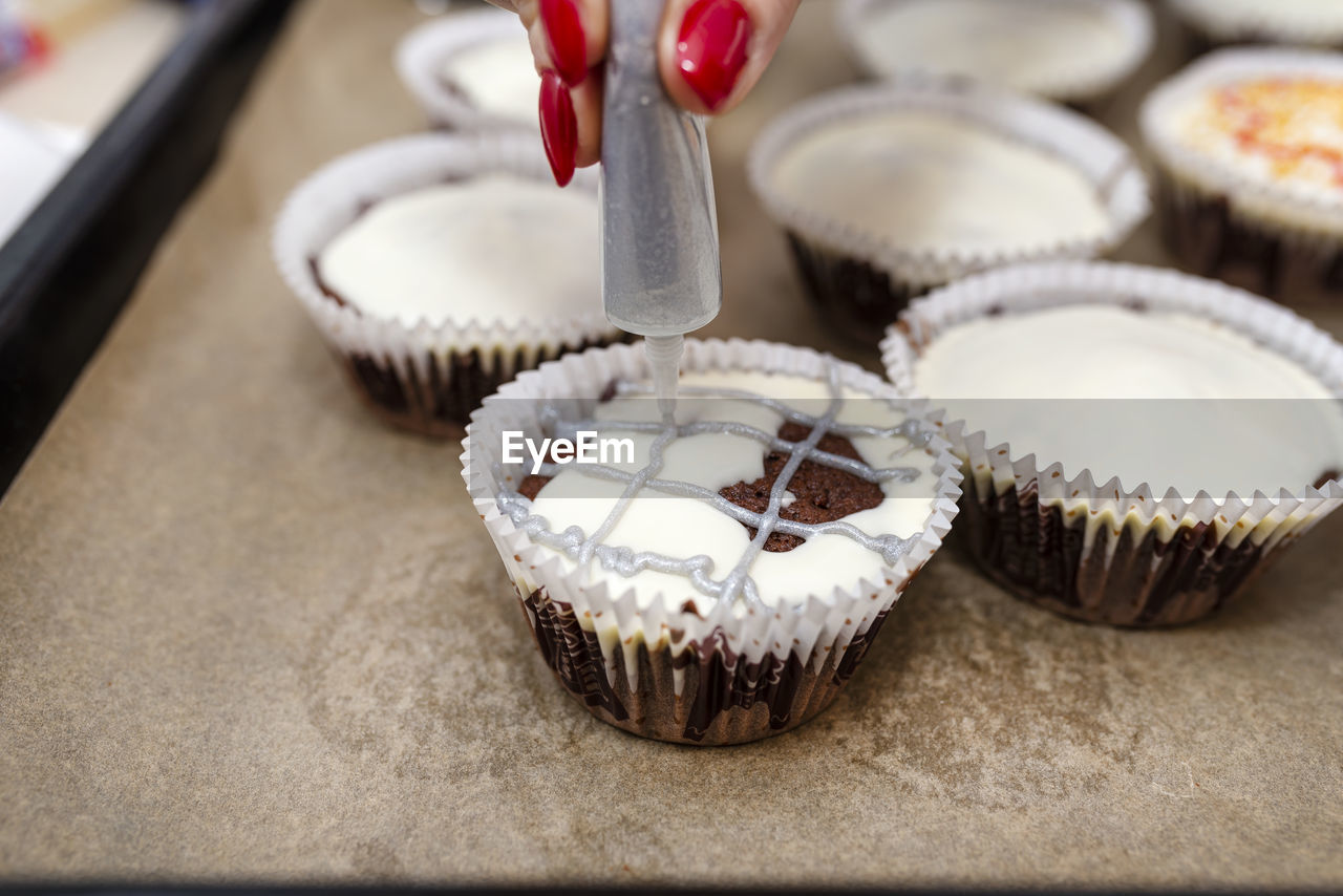 A woman squeezes colored frosting from a tube onto chocolate brown cupcakes covered white frosting.