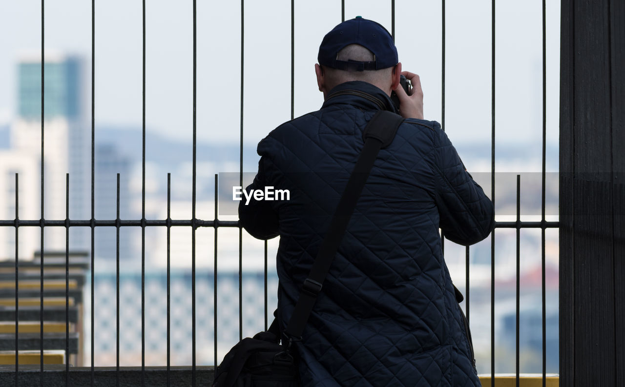 Rear view of man photographing with camera while standing by metal fence in city