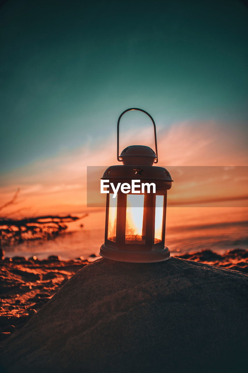 Close-up of old-fashioned lantern on rock at beach against sky during sunset