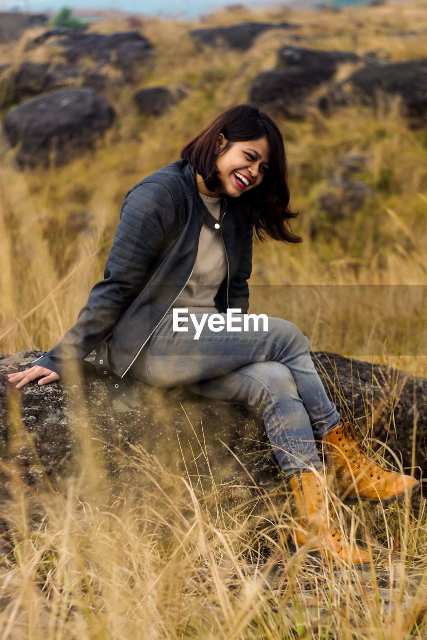 Full length of cheerful young woman sitting on rock at farm