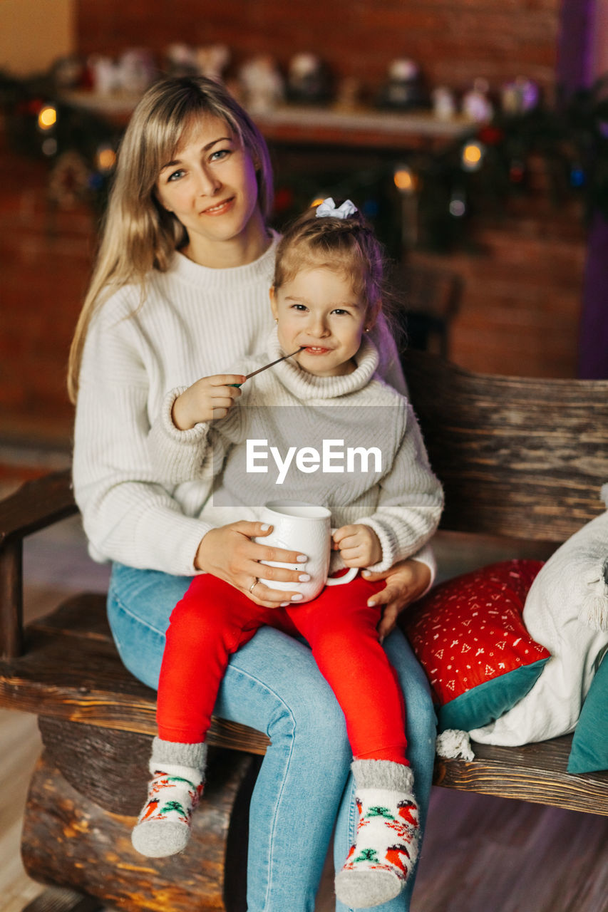 Portrait of a happy little girl sitting on her mother's lap and drinking hot chocolate on christmas