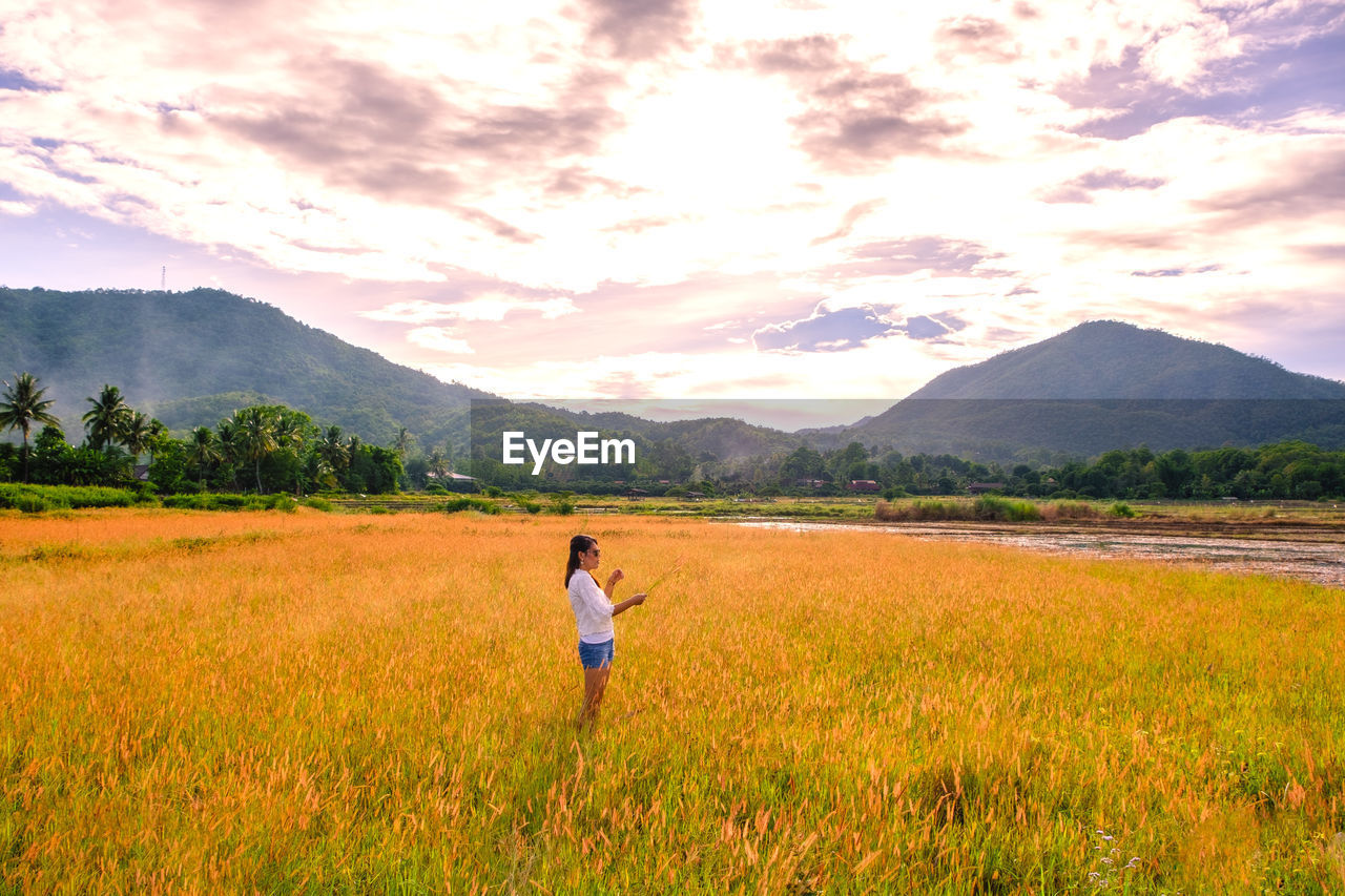 Side view of woman standing at farm against sky during sunset