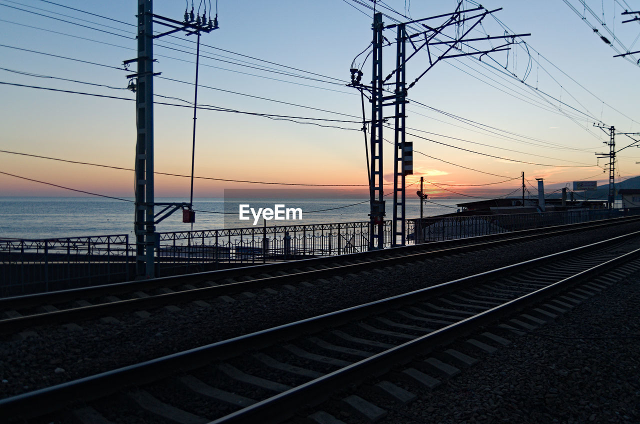 sky, sunset, transportation, electricity, cable, railroad track, track, rail transportation, line, power line, technology, electricity pylon, nature, power supply, mode of transportation, architecture, water, travel, sea, power generation, silhouette, vehicle, no people, transport, outdoors, railway, built structure, public transportation, beauty in nature, travel destinations, scenics - nature, city, evening, dusk, horizon, sun, twilight, sunlight, cloud, railroad station platform, train, tranquility, electrical supply, overhead power line, clear sky
