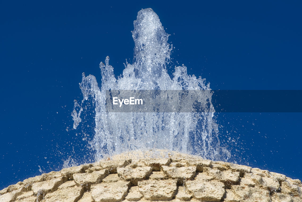 LOW ANGLE VIEW OF WAVES SPLASHING ON ROCKS AGAINST BLUE SKY