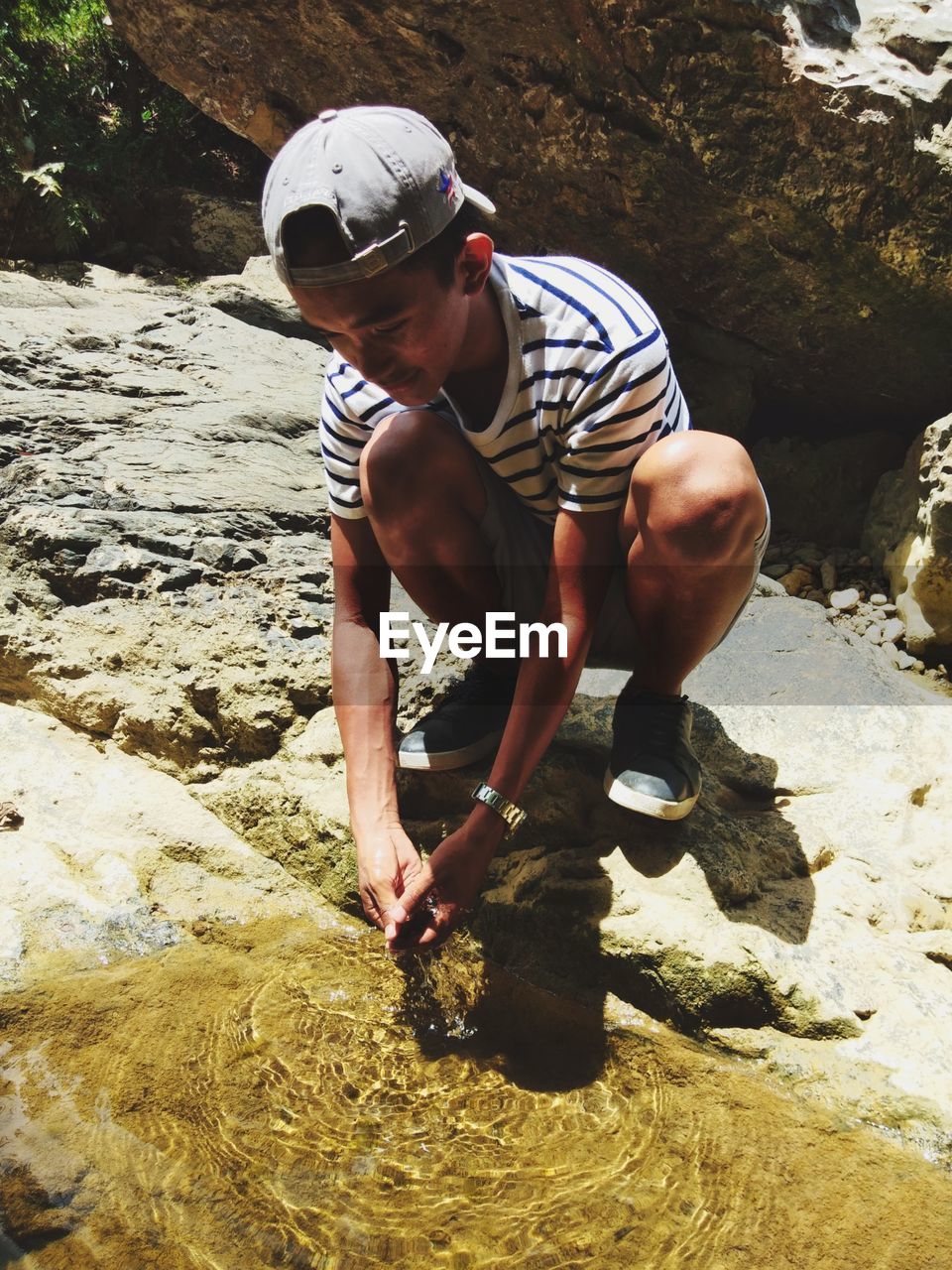 Full length of young man washing hands in water while crouching on rock during sunny day